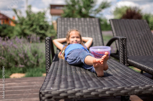 A cute little girl relaxing on the chaise longue at green garden outside, home feeling