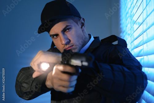 Professional security guard with flashlight and gun near window in dark room