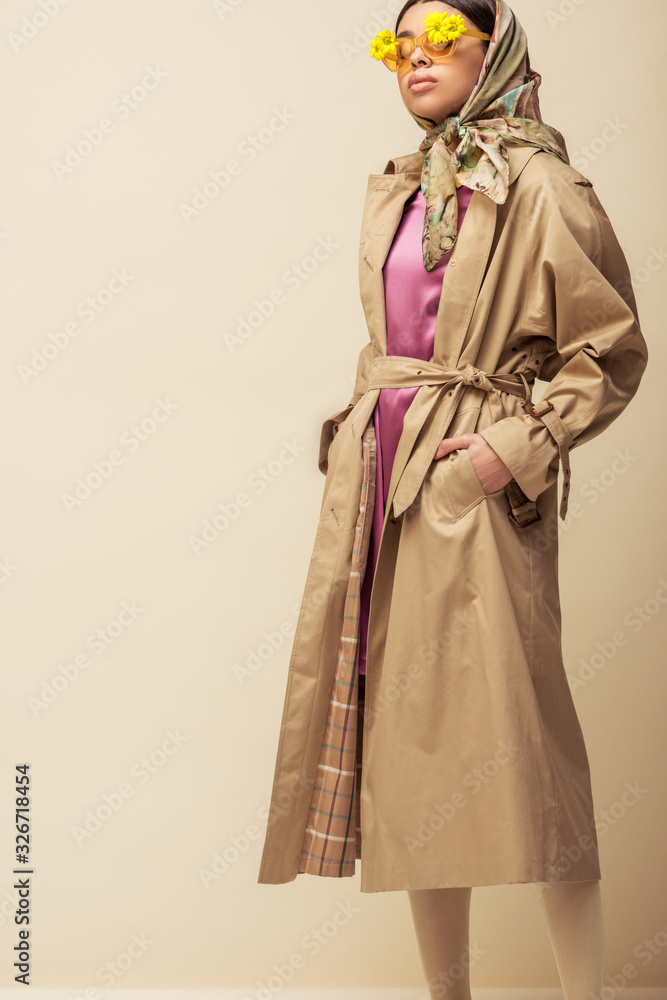 Fototapeta premium stylish african american girl in sunglasses with flowers, head scarf and trench coat standing with hands in pockets on beige