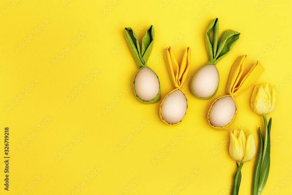 Funny Easter concept. Yellow flowers tulip and wooden easter egg with fabric ears as fun bunny on yellow background. Flat lay, top view.