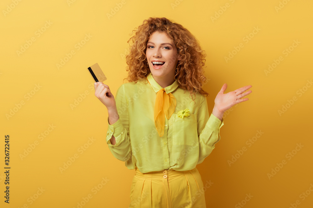 excited redhead woman holding credit card and gesturing on yellow excited redhead woman holding credit card and gesturing on yellow