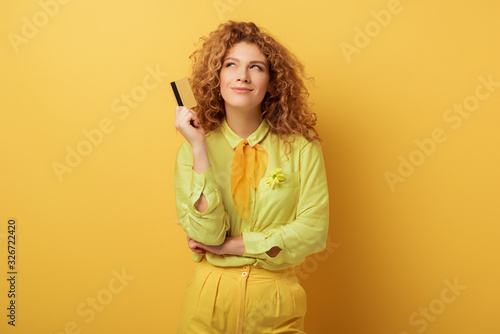 smiling redhead girl holding credit card while thinking on yellow