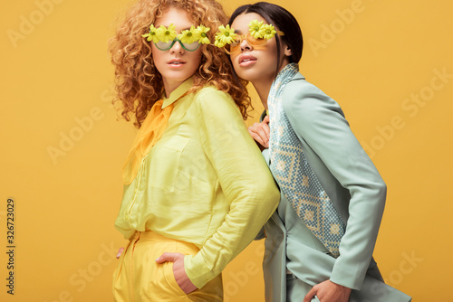 attractive multicultural women in sunglasses with flowers standing with hands in pockets isolated on yellow