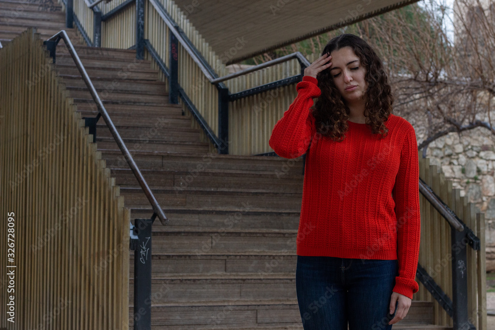 Portrait of caucasian young woman model with headache, in the park, orange sweater and jeans, long curly hair. Place for your text in copy space. Portrait of caucasian young woman model with headache, in the park, orange sweater and jeans, long curly hair. Place for your text in copy space.