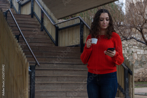 Portrait of attractive caucasian young woman model drinking coffee and looking at phone, in the park, orange sweater and jeans, long curly hair. Place for your text in copy space.