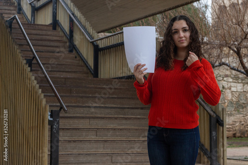pretty young woman model holding white empty paper, in the park, orange sweater and jeans, long curly hair. Place for your text in copy space.