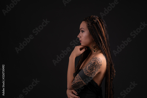 Shot of a young woman with a tattoo on her arms and with braids against dark background with space in the studio