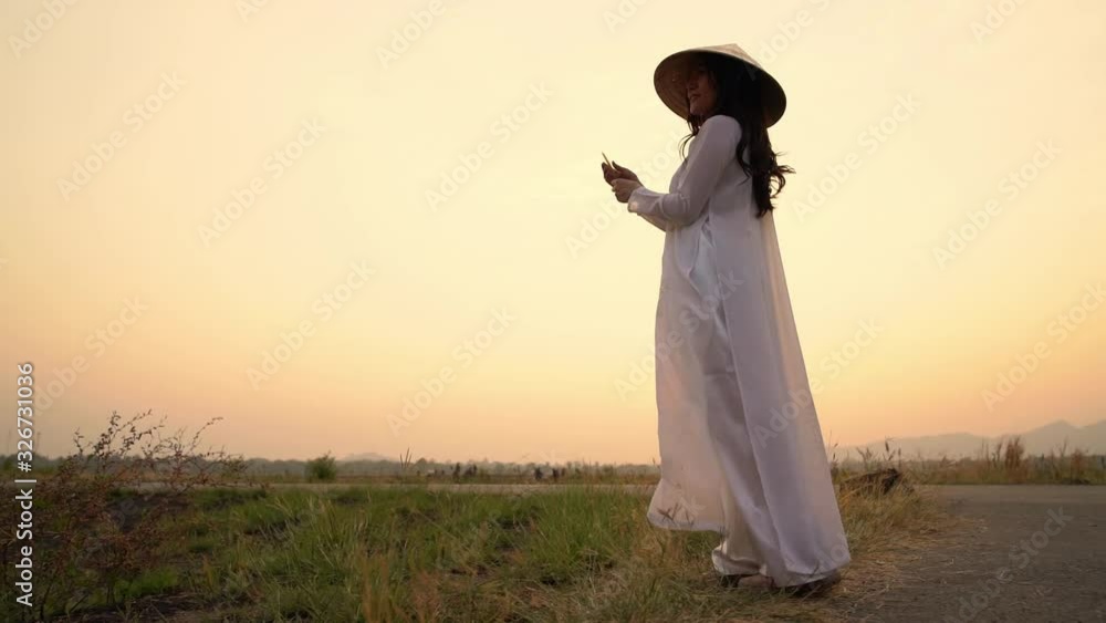 The silhouette scene of a Vietnamese woman in traditional costume holding grass flower little swaying from the wind in the autumn sunset, natural field flower, relax naturally atmosphere. The silhouette scene of a Vietnamese woman in traditional costume holding grass flower little swaying from the wind in the autumn sunset, natural field flower, relax naturally atmosphere.