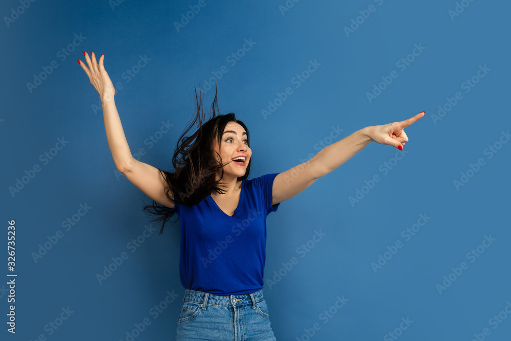Pointing on. Beautiful caucasian woman's portrait isolated on blue studio background. Beautiful female brunette model in casual style. Concept of human emotions, facial expression, sales, ad