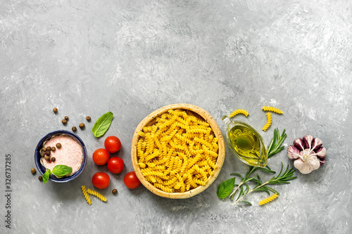 Border with ingredients for cooking pasta with tomato sauce. Fusilli, tomatoes, basil, olive oil, pink salt, pepper, rosemary and garlic on a gray concrete background. Top view, flat lay,copy space.