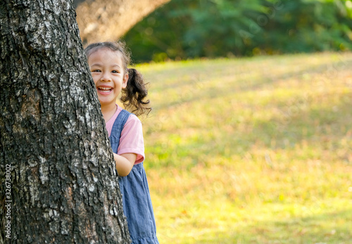 Fototapeta Naklejka Na Ścianę i Meble -  Portrait of little Asian girl play peekaboo by hiding back of tree and smile in park or garden.