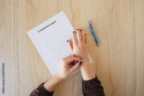 Student girl hand with bracelet and love word drawing a heart on empty examination paper sheet on the school desk. First love during university exam. Concept