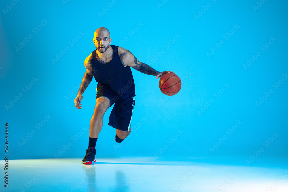 Young basketball player of team wearing sportwear training, practicing in action, motion isolated on blue background in neon light. Concept of sport, movement, energy and dynamic, healthy lifestyle.