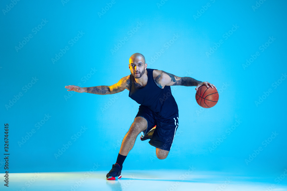 Young basketball player of team wearing sportwear training, practicing in action, motion isolated on blue background in neon light. Concept of sport, movement, energy and dynamic, healthy lifestyle. Young basketball player of team wearing sportwear training, practicing in action, motion isolated on blue background in neon light. Concept of sport, movement, energy and dynamic, healthy lifestyle.