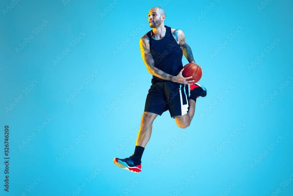 In jump. Young basketball player of team wearing sportwear training, practicing in action, motion on blue background in neon light. Concept of sport, movement, energy and dynamic, healthy lifestyle.