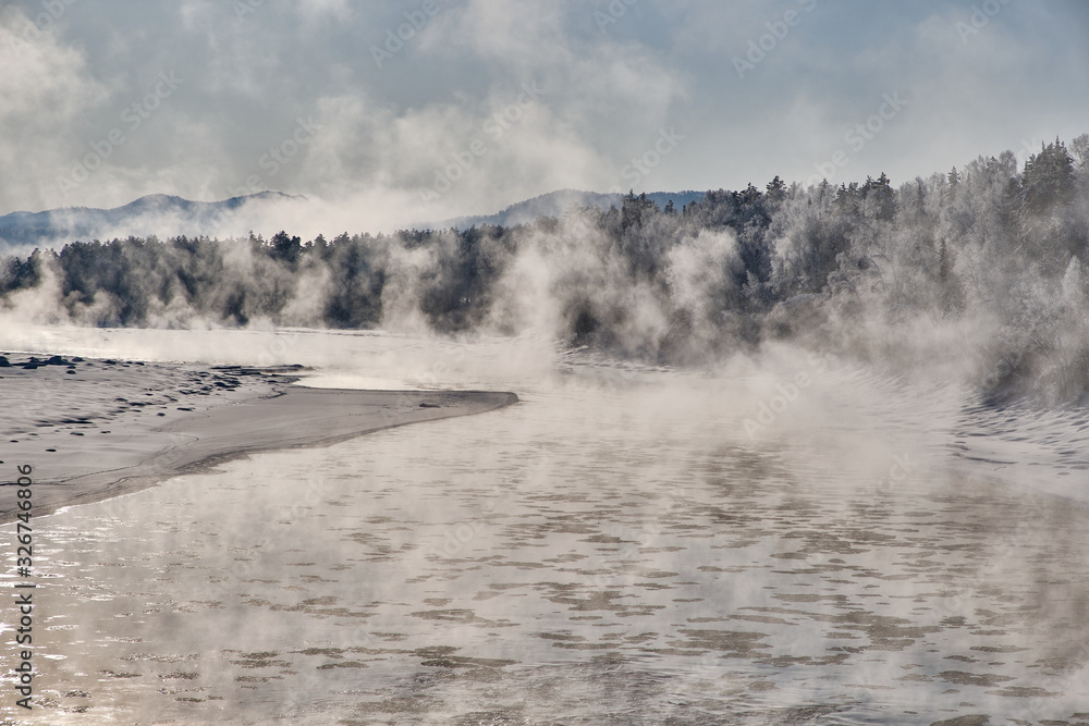 Winter landscape. Not freezing river with snowy trees along the banks. Steam rises from the winter mountain river against the blue sky. Morning back light. Russia. Altai Republic. Winter landscape. Not freezing river with snowy trees along the banks. Steam rises from the winter mountain river against the blue sky. Morning back light. Russia. Altai Republic.