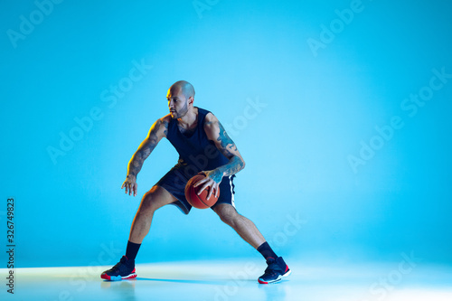 Young basketball player of team wearing sportwear training, practicing in action, motion isolated on blue background in neon light. Concept of sport, movement, energy and dynamic, healthy lifestyle. © master1305
