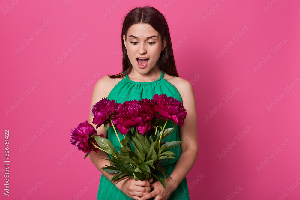 Beautiful cheerful girl stands isolated over pink studio background, holding bouquet of burgundy peony flowers, having astonished facial expression, poses with widely opened mouth. Surprise at 8 March Beautiful cheerful girl stands isolated over pink studio background, holding bouquet of burgundy peony flowers, having astonished facial expression, poses with widely opened mouth. Surprise at 8 March