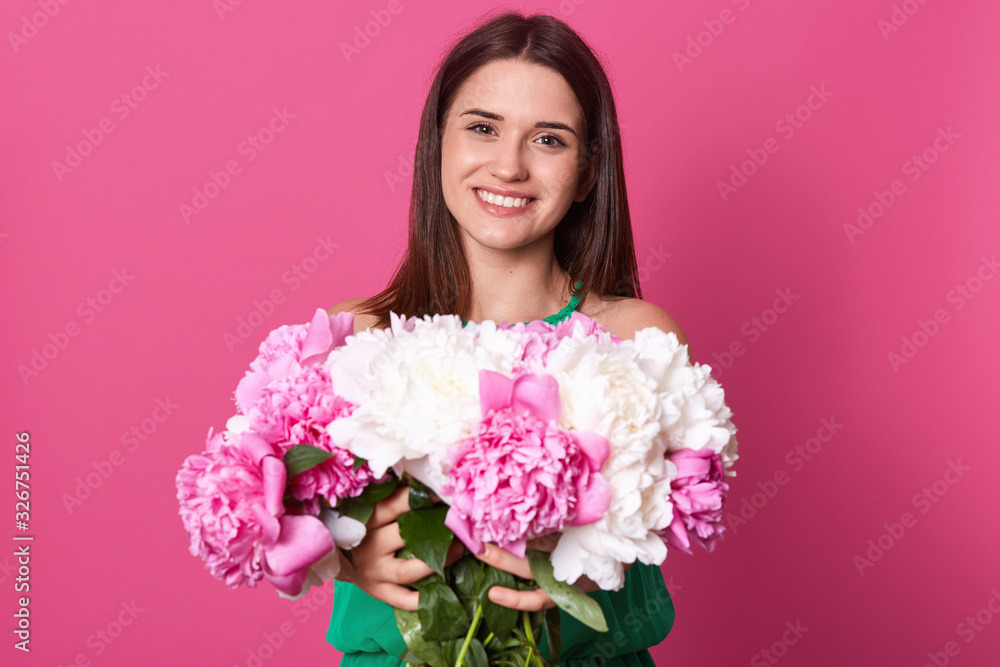 Beautiful joyful dark haired woman with gentle smile, looks with cheerful expression directly at camera, recieving big bouquet of flowers, isolated over rosy studio wall. Women's day concept. Beautiful joyful dark haired woman with gentle smile, looks with cheerful expression directly at camera, recieving big bouquet of flowers, isolated over rosy studio wall. Women's day concept.