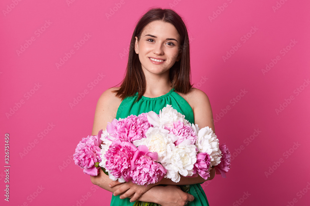 Studio shot of beautiful brunette girl holding with white and rose peonies in hands, attractive woman looking at camera, female embraces flowers, dressed green sundress, posing against pink background Studio shot of beautiful brunette girl holding with white and rose peonies in hands, attractive woman looking at camera, female embraces flowers, dressed green sundress, posing against pink background