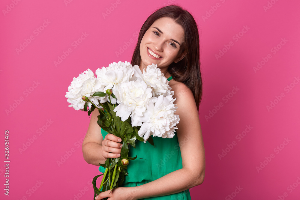 Brunette cute female with bouquet of white peonies in hands, posing with charming, girl in gren sundress. Dark haired lady holding her present from boyfriend, looking at camera with happy expression.
