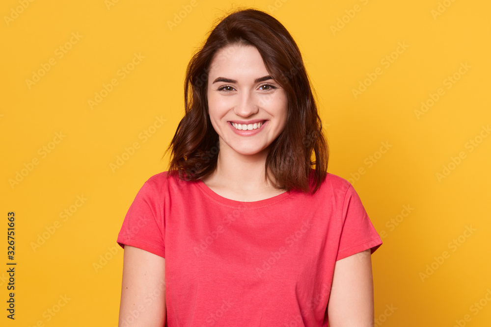 Picture of Caucasian brunette woman posing isolated over yellow background, dark haired lady looks smiling directly at camera, looking happy, lady wearing red t shirt. People emotions concept. Picture of Caucasian brunette woman posing isolated over yellow background, dark haired lady looks smiling directly at camera, looking happy, lady wearing red t shirt. People emotions concept.