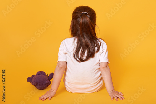Φωτογραφία Rear view of little fair haired child sitting on the floor, putting hands aback, being punished, spending time alone, having brown teddy bear nearby, wearing white t shirt