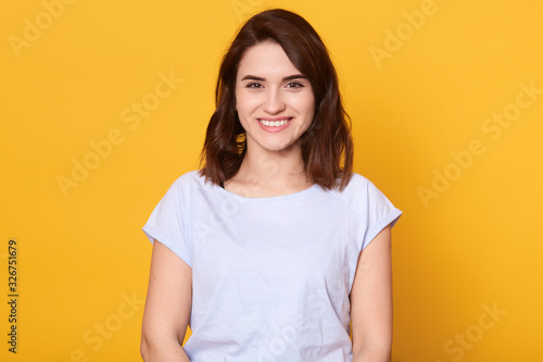 Head shot portrait of young woman having wide white smile, wearing white t shirt, lady looking at camera, pretty female posing isolated over bright yellow background, girl expressing happyness.