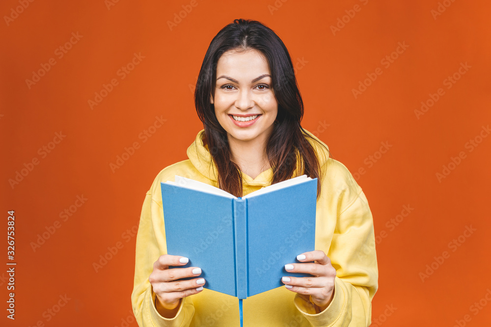 Beautiful young woman wearing casual standing isolated over orange background, reading a book. Beautiful young woman wearing casual standing isolated over orange background, reading a book.