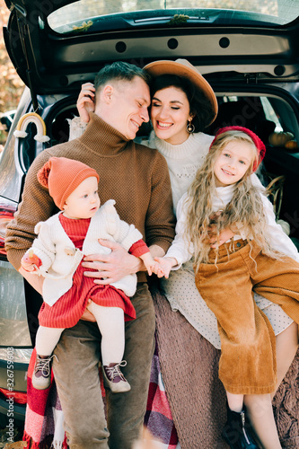 a happy family of four people in an autumn pine forest sit in the cozy trunk of their car covered with blankets