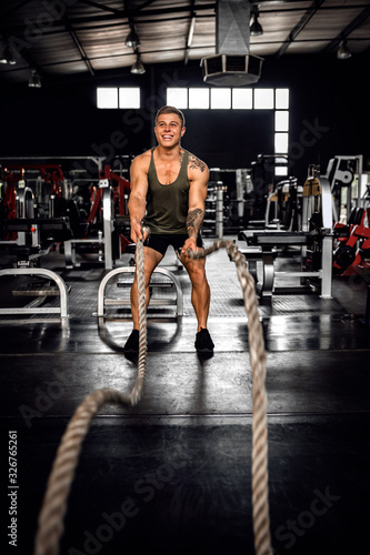 Smiling young man exercising with battle ropes in a gym