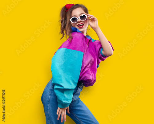 Cool teenager. Fashionable DJ girl in colorful trendy jacket and vintage retro sunglasses enjoys style of 80s � 90s vibes. Teenager Girl at disco party. Young fashion model on yellow color background.