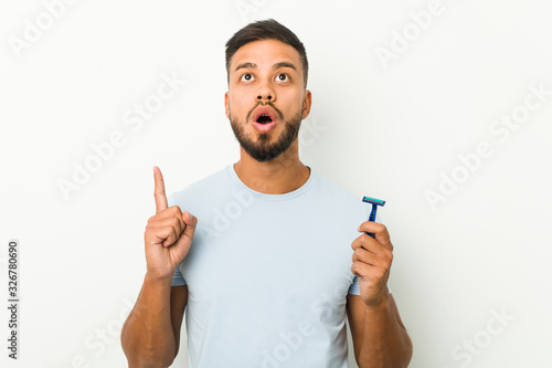 Young south-asian man holding a razor blade pointing upside with opened mouth.