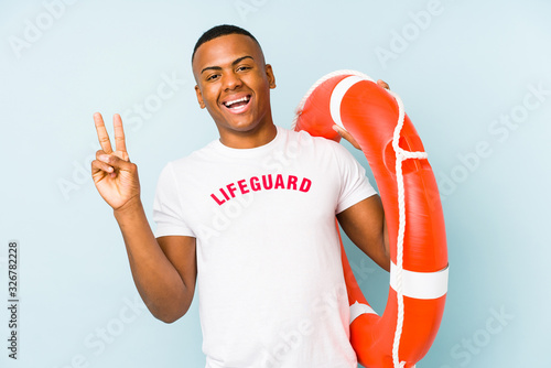 Young latin life guard isolated on blue background joyful and carefree showing a peace symbol with fingers. © Asier