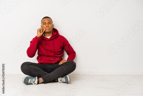 Young latin man sitting on the floor isolated with fingers on lips keeping a secret. © Asier