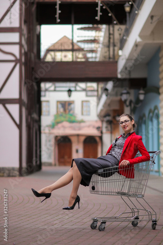 Beautiful cute young businesswoman in red jacket and formal suit and high-heeled shoes and glasses sits in shopping cart on blurred background of buildings