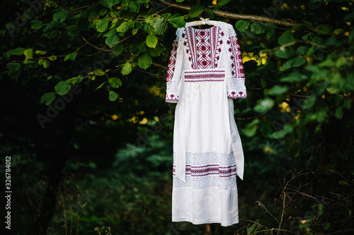 White, red wedding dress for bride on hanger hanging on tree branch in garden outdoors. Photo made before ceremony of marriage. Ukrainian style: embroidered clothes on nature. Ethnic national costume. © Serhii