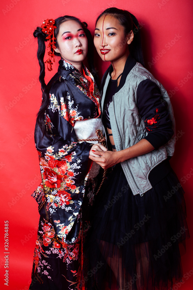 two pretty geisha girls friends: modern asian woman and traditional wearing kimono posing cheerful on red background