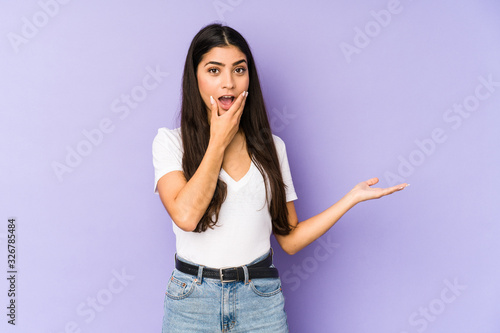Young indian woman isolated on purple background holds copy space on a palm, keep hand over cheek. Amazed and delighted.