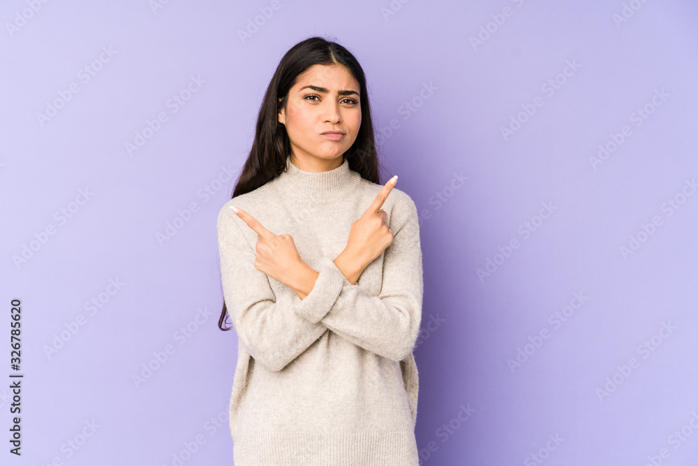 Young indian woman isolated on purple background points sideways, is trying to choose between two options. Young indian woman isolated on purple background points sideways, is trying to choose between two options.