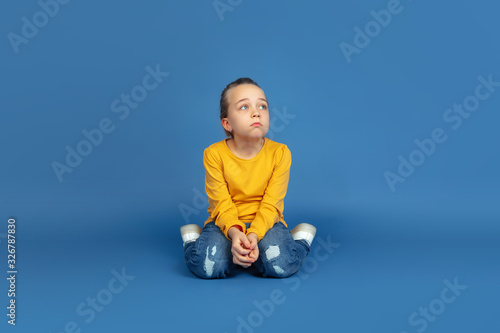 Portrait of sad little girl sitting isolated on blue studio background. How it feels to be autist. Modern problems, new vision of social issues. Concept of autism, childhood, healthcare, medicine. photo