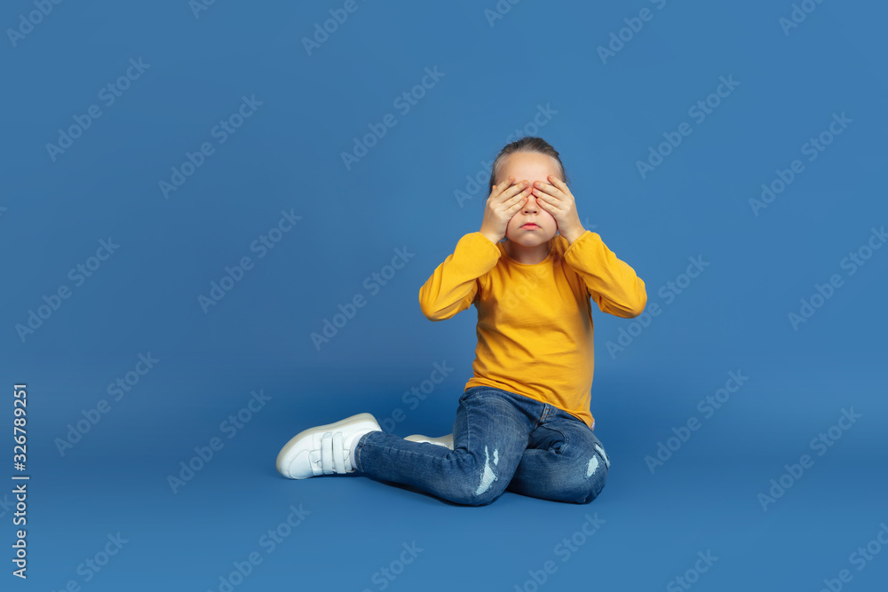 Portrait of sad little girl sitting isolated on blue studio background. How it feels to be autist. Modern problems, new vision of social issues. Concept of autism, childhood, healthcare, medicine. Portrait of sad little girl sitting isolated on blue studio background. How it feels to be autist. Modern problems, new vision of social issues. Concept of autism, childhood, healthcare, medicine.