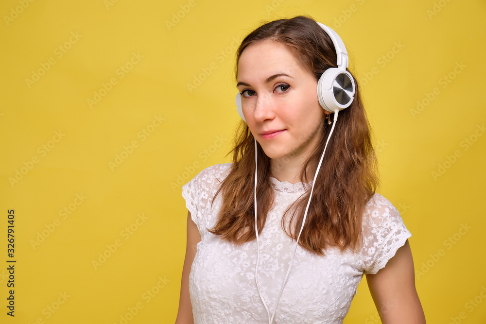A girl in summer clothes stands on a yellow background, listens to music in white headphones and looks at the camera. Portrait.