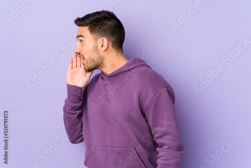 Young caucasian man isolated on purple background is saying a secret hot braking news and looking aside