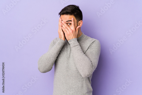 Young caucasian man isolated on purple background blink through fingers frightened and nervous.