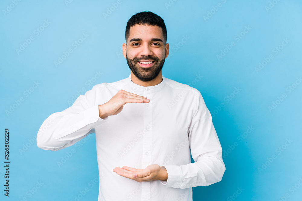 Young latin man against a blue background isolated holding something with both hands, product presentation. Young latin man against a blue background isolated holding something with both hands, product presentation.