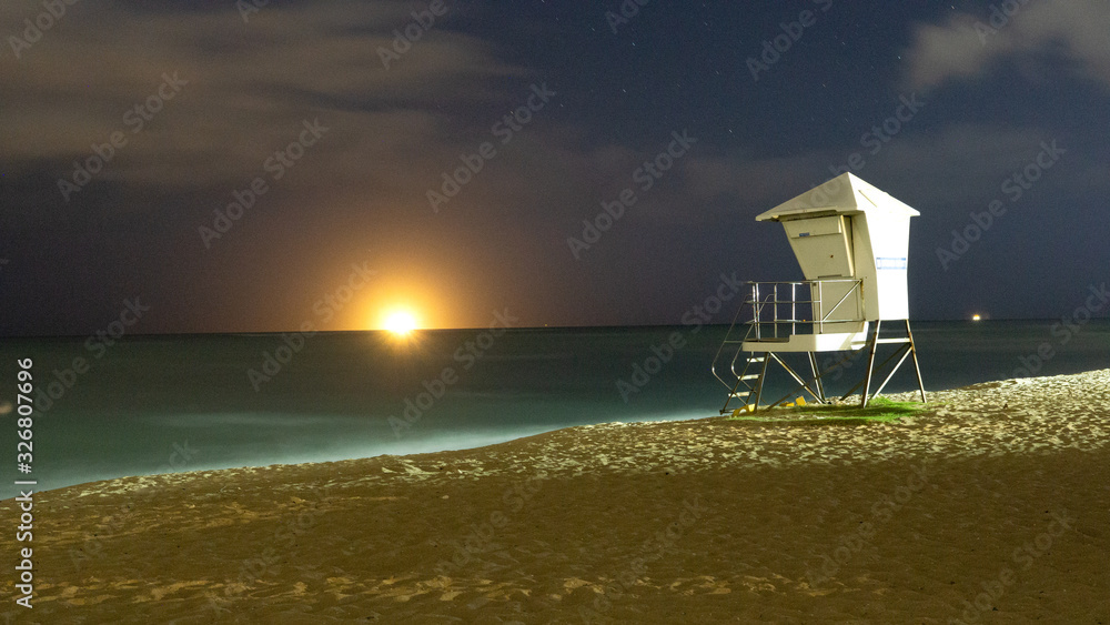 Lifeguard stand at White Plains Beach Hawaii Oahu at night lighted by street lights with an Oil Tanker on the horizon with lights that make it look like the sun