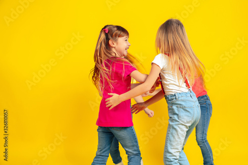 Running. Happy children playing and having fun together on yellow studio background. Caucasian kids in bright clothes looks playful, laughting, smiling. Concept of education, childhood, emotions.