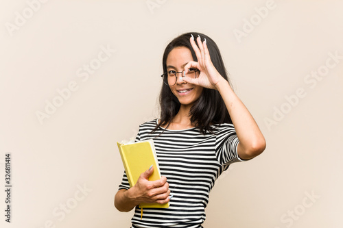 Young asian woman student holding a book excited keeping ok gesture on eye.