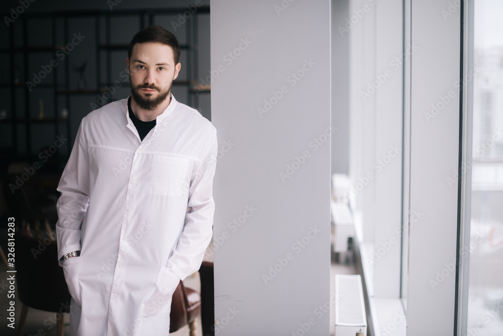 Confident young male doctor. Caucasian doctor with beard stands by the window and looking at camera. Male doctor posing in modern meeting office room of the hospital. Concept of medical work.